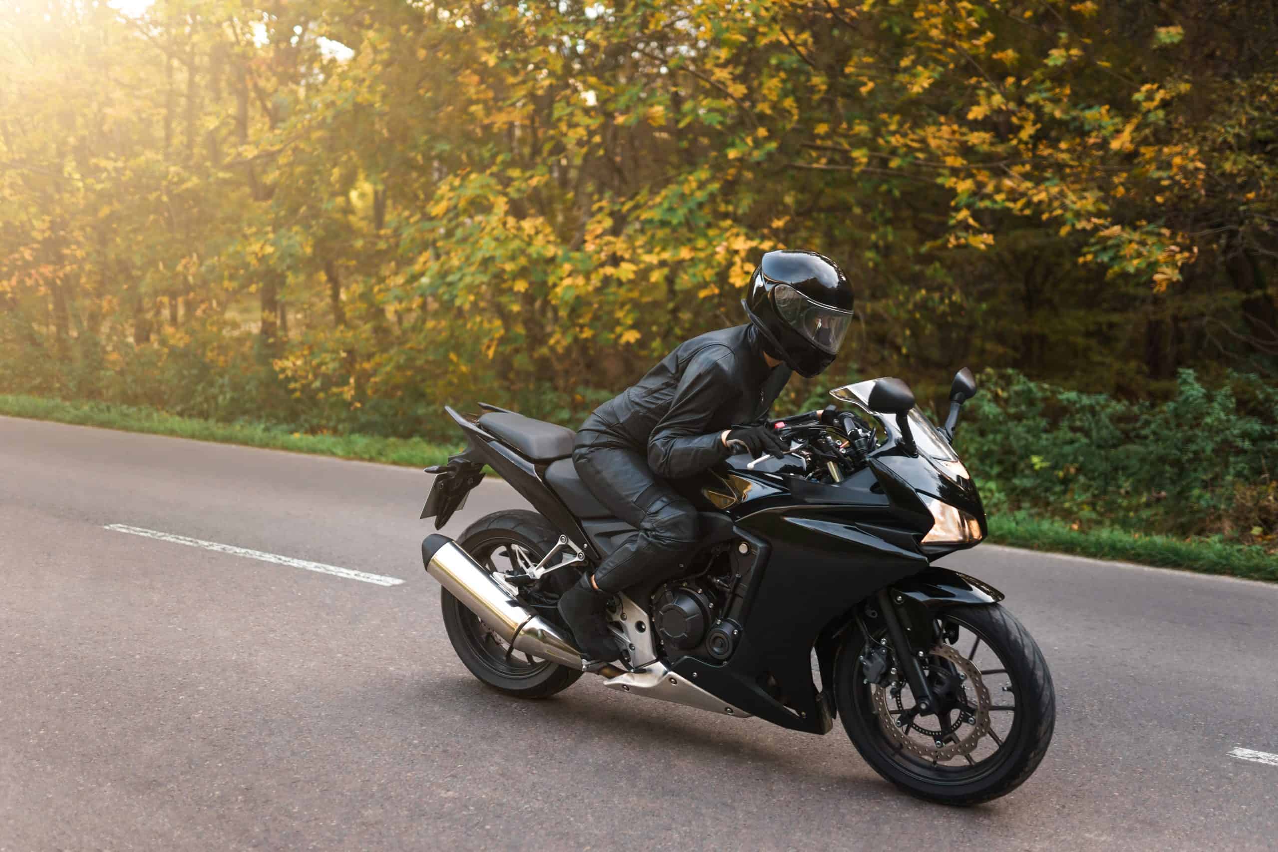 A person in black motorcycle gear rides a black sport bike on a paved road with trees and greenery in the background.