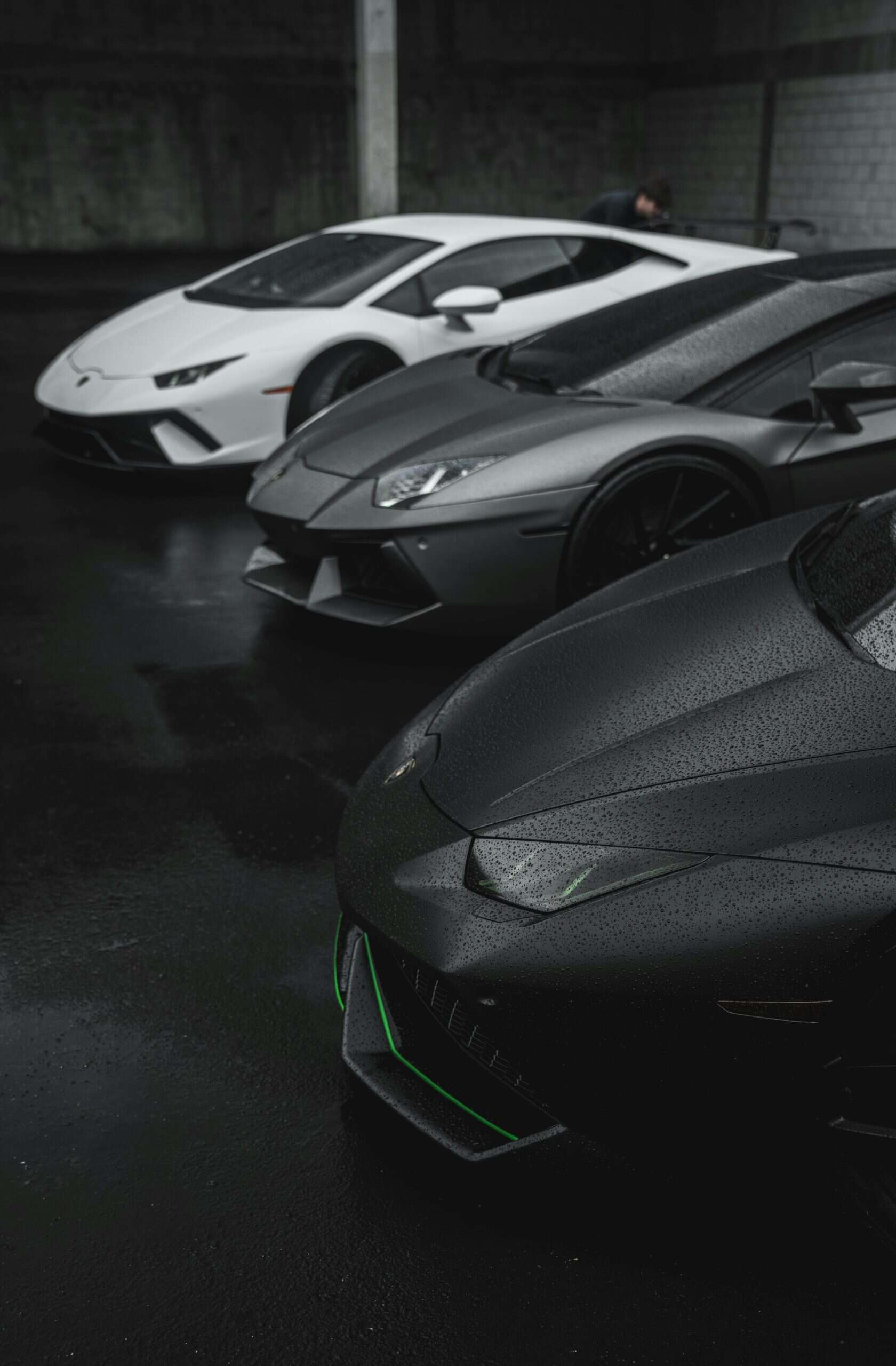Three Lamborghini sports cars in black, gray, and white are parked in a row on a wet, dark pavement in an industrial setting.