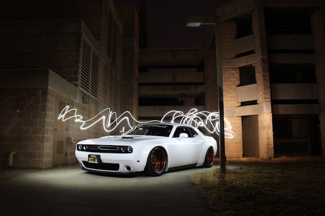 A white Dodge Challenger parked outdoors at night near a building, with white light trails streaking around the car.